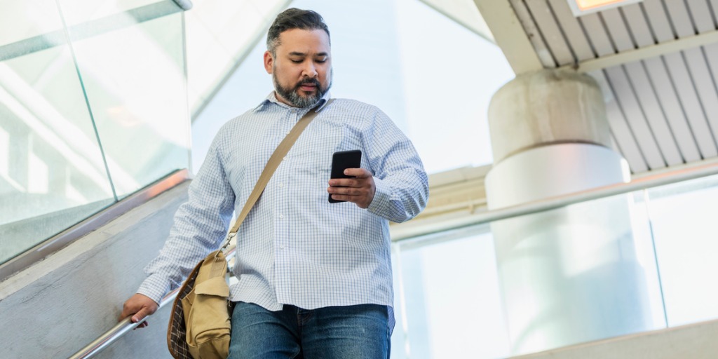 A man in his 30s wearing a button down shirt, walking down stairs outside a building, looking down at his mobile phone.