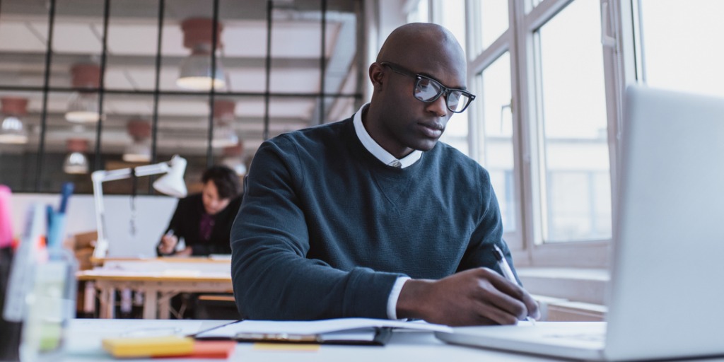 A man writing notes while working on a laptop in a modern office. A man writing notes while working on a laptop in a modern office.