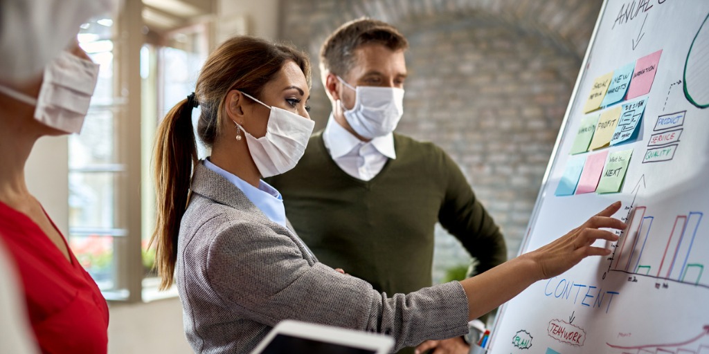 Businesswoman and her coworkers wearing protective face masks while making new business strategy on whiteboard during coronavirus epidemic.