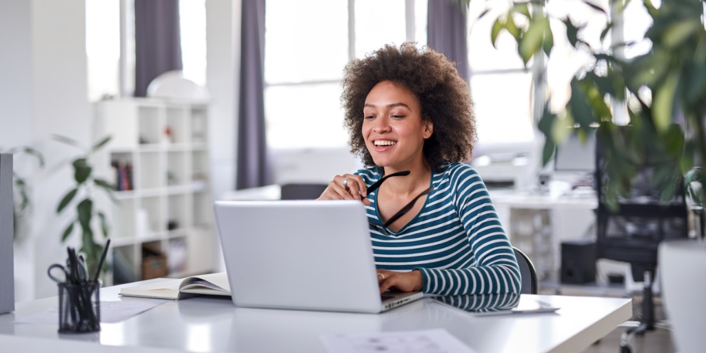 Smiling woman holding eyeglasses and looking at laptop while sitting and working in a modern office