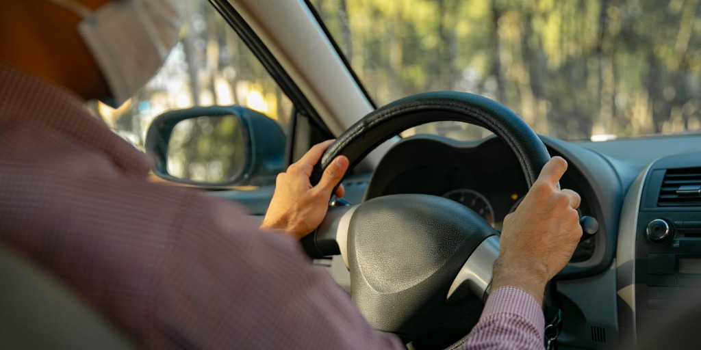 Adult man wearing a red shirt and a mask, driving a car. 