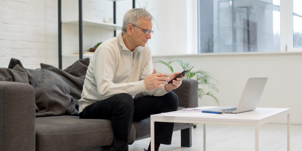 Busy older male working with smartphone and laptop while staying in living room