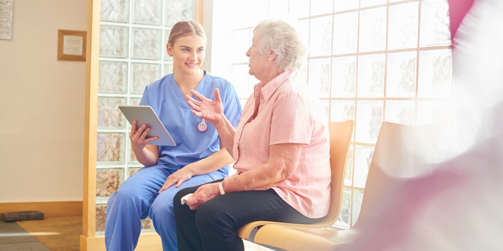 A female nurse sits in doctor’s waiting room with senior female patient taking notes and updating her medical records