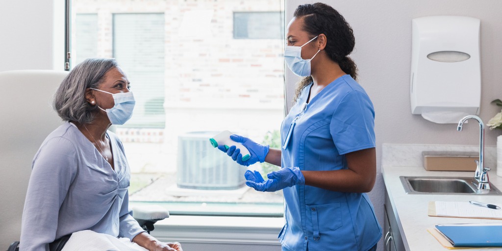 A nurse gestures while talking with a female patient. The nurse is holding an infrared thermometer.