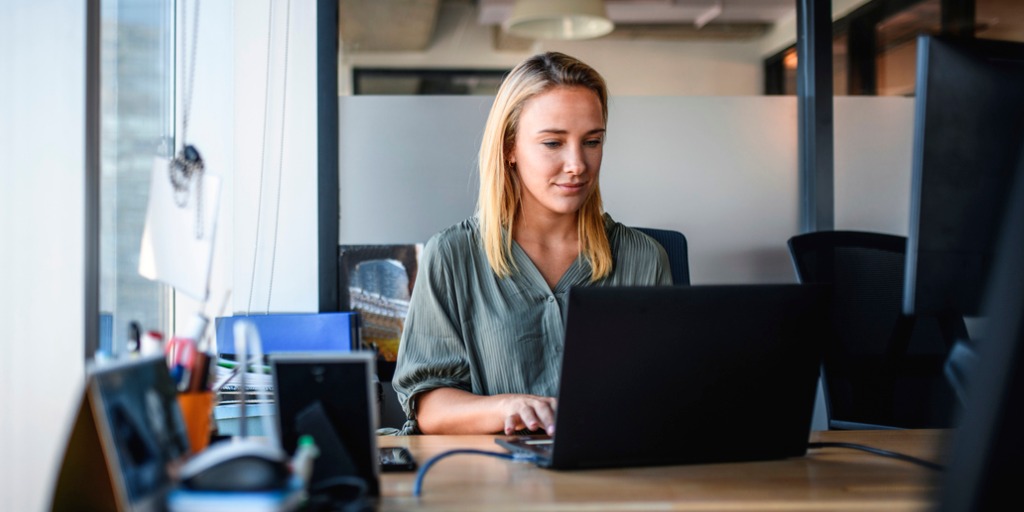 Desk-level perspective of blonde woman typing on laptop in office with window view.