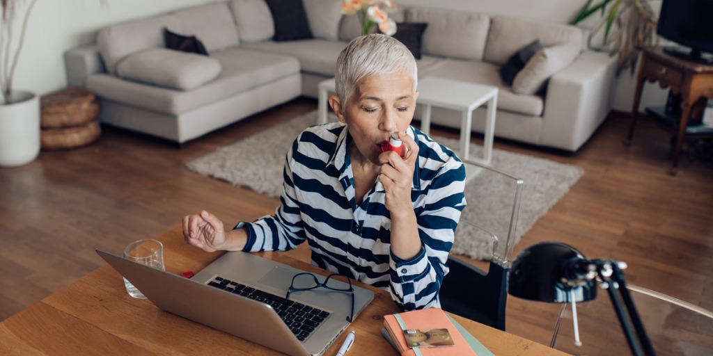 Mature woman using asthma inhaler while working on a laptop at home.