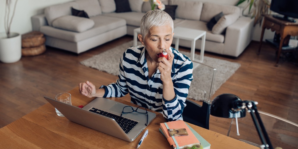 Mature woman using asthma inhaler while working on a laptop at home.