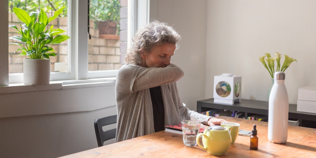 Middle aged woman sitting at table coughing into elbow Middle aged woman sitting at table coughing into elbow