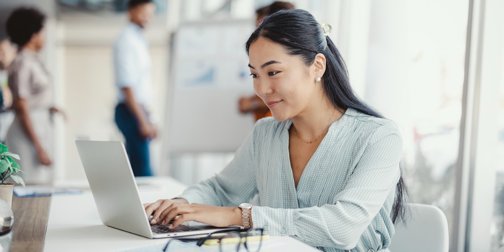 Businesswoman busy working on laptop computer at office with colleagues in the background.