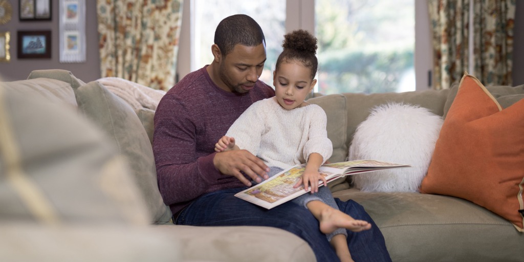 An African-American pre-school age girl sits on dad's lap in the living room and reads a book. She is pointing out her favorite parts and making sure he's not skipping any pages...