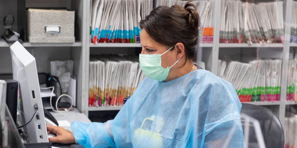 Receptionist at medical office wearing mask and gown for protection during the Covid-19 pandemic. She is working at the computer with patient files in the background.