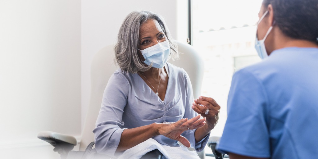 A senior woman, wearing a protective face mask, talks with a female nurse during a medical appointment.